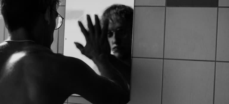 A shirtless man with glasses touching a mirror.