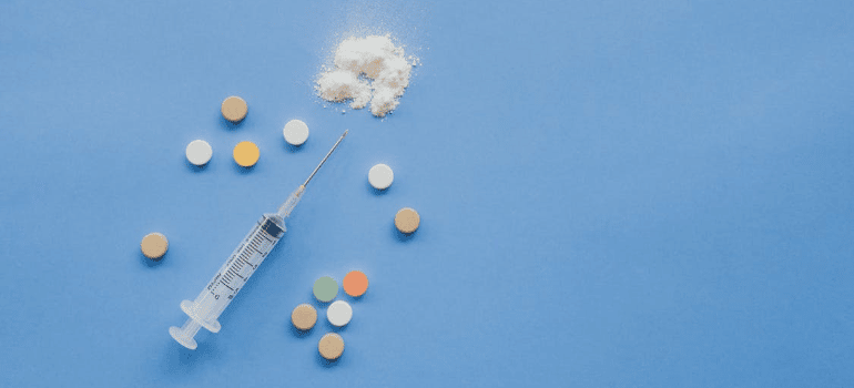 A syringe next to pills and white powder on a blue surface.