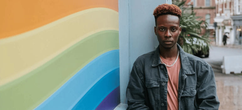 A black man leaning on a wall which features a rainbow flag graffiti.