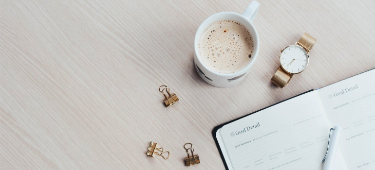 A planner book by a watch and a mug of coffee on a wooden surface.