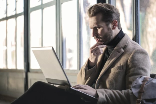 A man in a brown jacket using a laptop as he touches his chin while checking out alcohol rehab Athens OH