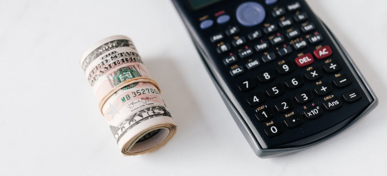 A roll of dollar bills next to a black calculator on a white surface.