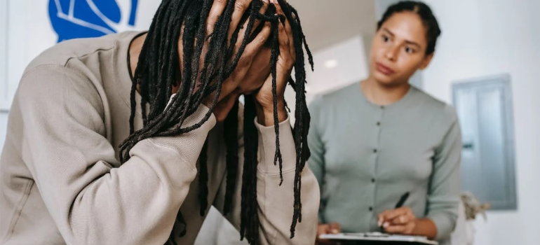 A psychotherapist in a long-sleeved shirt keeping notes as her patient holds his head in distress.