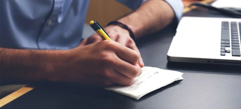 A close-up of a person writing on a paper by a laptop.