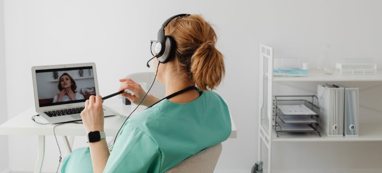 A clinician in a teal scrub suit on a video call with a patient on a laptop.