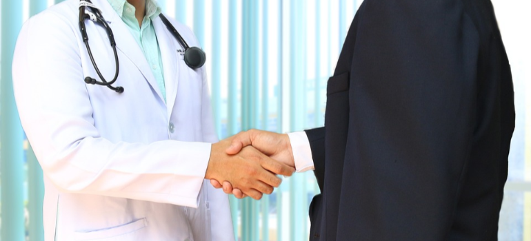 A doctor in a white suit and a patient in a black suit shaking hands.