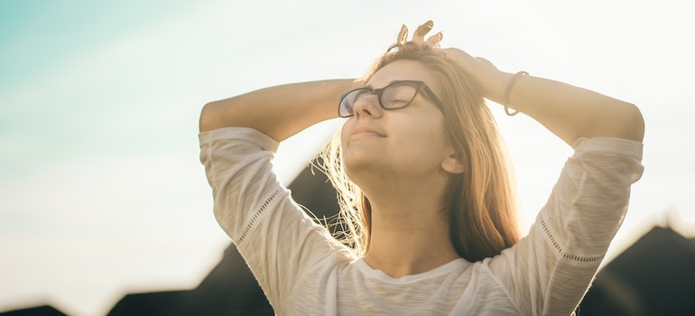 A happy woman standing in the sun and smiling with her arms behind her head.
