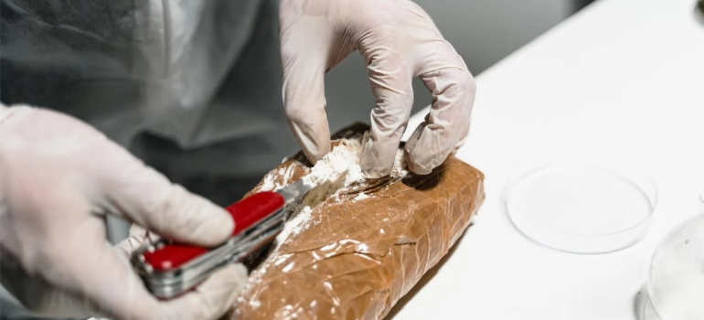 A person wearing surgical gloves removing white powder from a package with a knife.