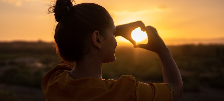trauma shapes addiction and healing but self-love can change that shown by a girl making a heart with her hands at sunset