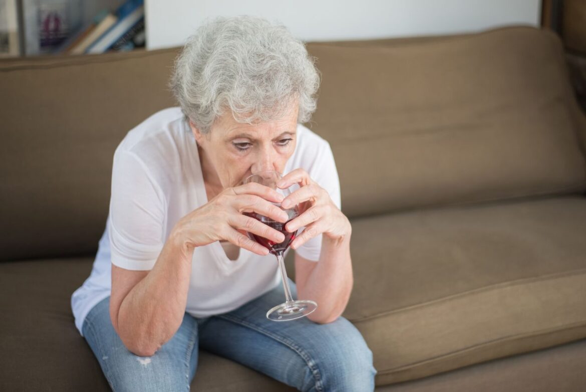 Elderly woman sitting on the couch and drinking a glass of wine.