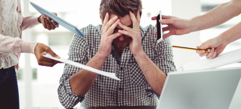 A man before a computer holds his hands while stressed while hands around him want to give him phones, papers, and pencils