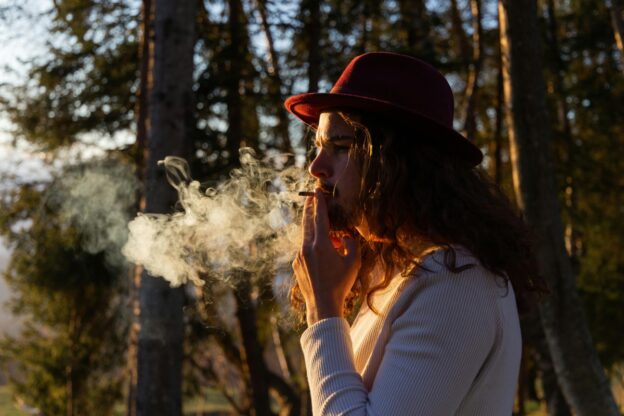 Man in white blouse and hat on his head, smoking cigarettes in the forest.