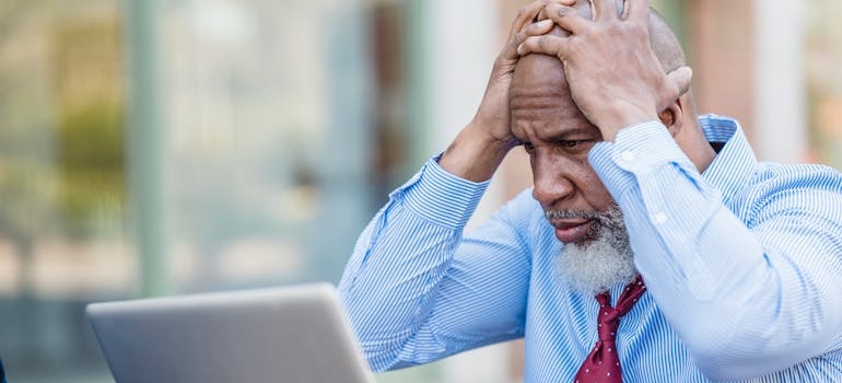 An older man holding his head and looking at a laptop.