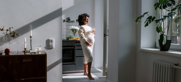 A pregnant woman is standing near the window holding a glass of water.