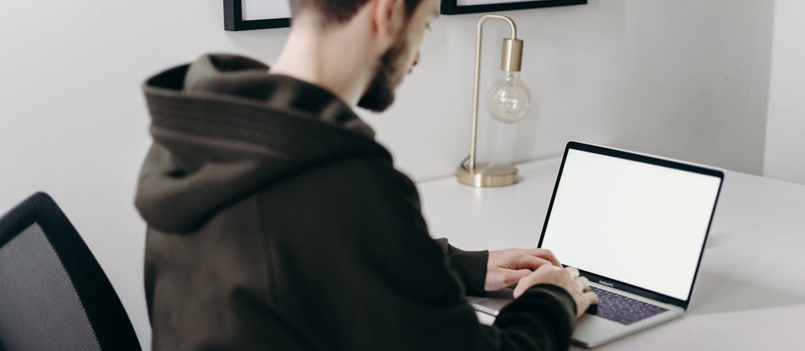 A man in a black hoodie is sitting and typing "FAQs about addiction treatment" on his laptop.