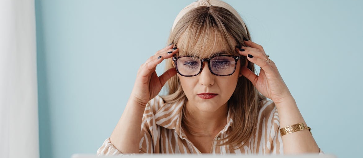 A woman in glasses is sitting and looking at her laptop, and wondering about FAQs about addiction treatment.
