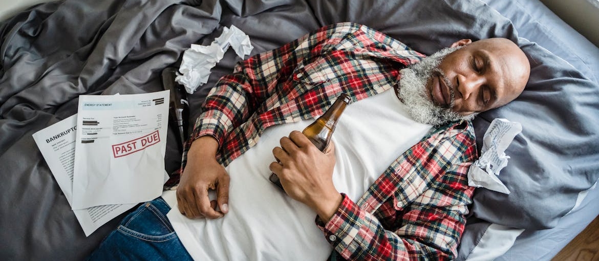 Man holding a beer bottle and sleeping surrounded by unpaid bills.