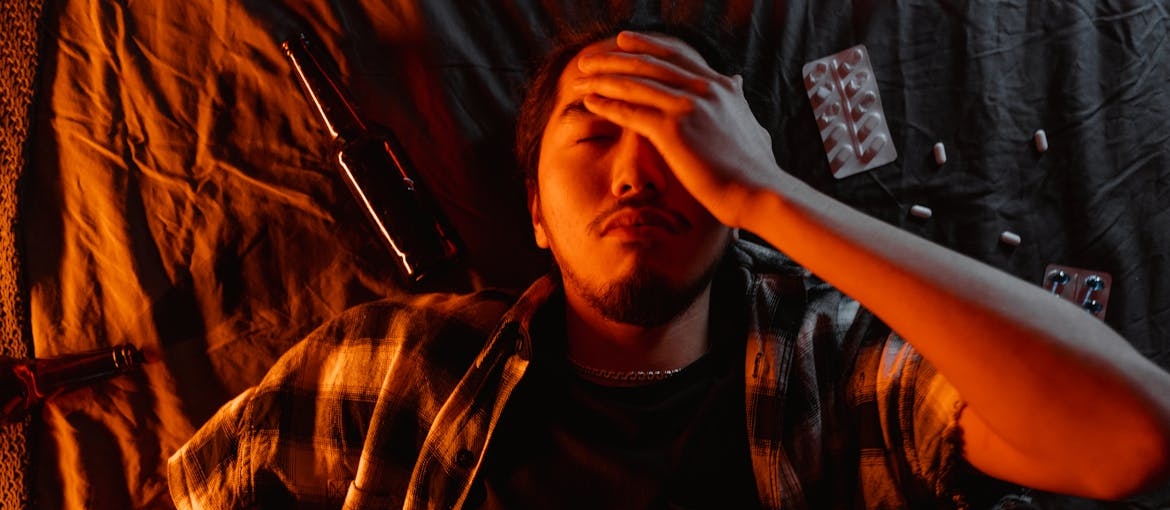 Man lying in bed surrounded by pills and beer bottles.