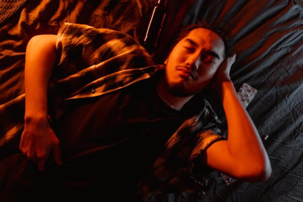 Man lying on the bed, surrounded by empty bottles of beer and pills while feeling the signs and symptoms of Adderall abuse.