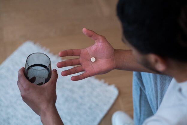 Man sitting on the couch holding a white pill and a glass of water.