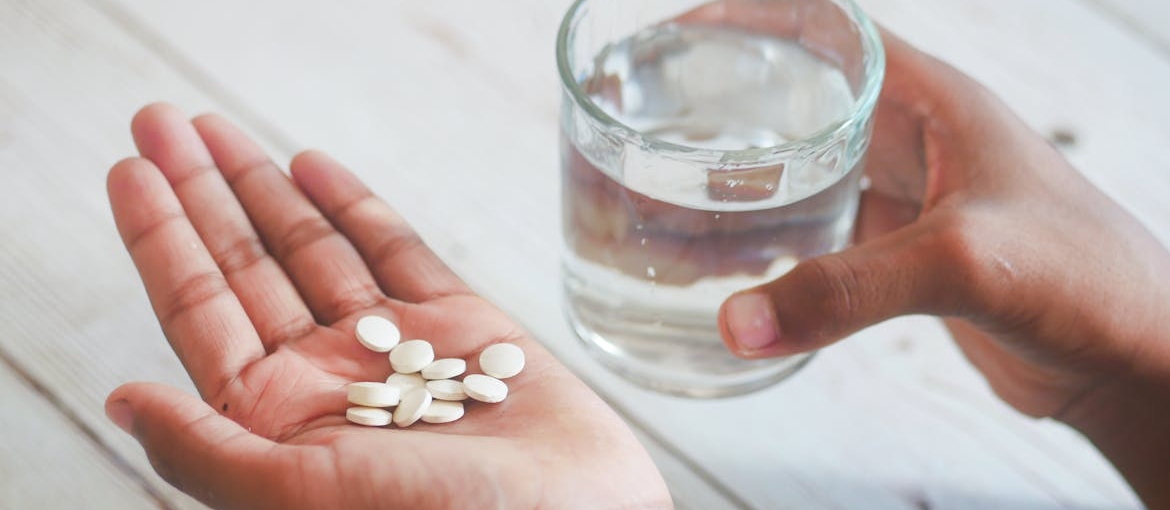 Person holding white pills in the palm of their hand and holding a glass of water.